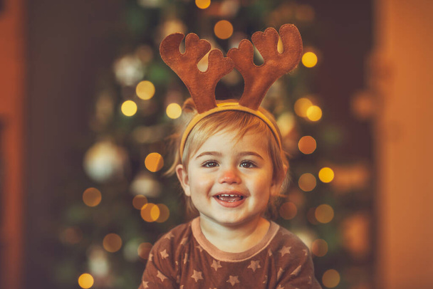 Portrait of a cheerful laughing baby wearing Rudolphs reindeer costume, with pleasure spending Christmas at home near beautiful glowing Christmas tree - Fotó, kép