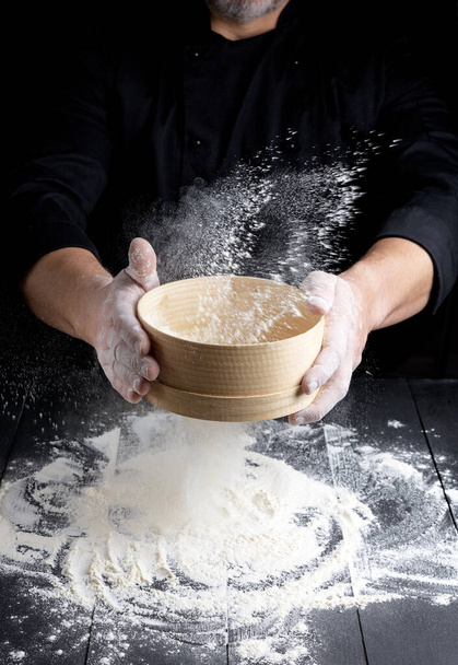 丸みを帯びた木のふるい男の手に小麦粉、シェフは黒い木製のテーブルの上に白い小麦粉をふるい - 写真・画像