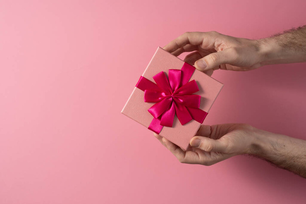 Valentine's Day celebration concept. A nice gift from a loved one. Box with a bow in male hands on a delicate pink background. Copy space. Flat lay. - Фото, изображение