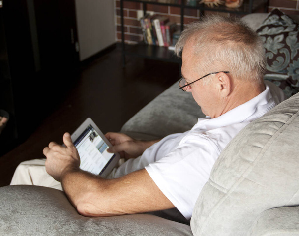 muž ve věku používá digitální přístroj, zatímco sedí na pohovce v interiéru. - Fotografie, Obrázek