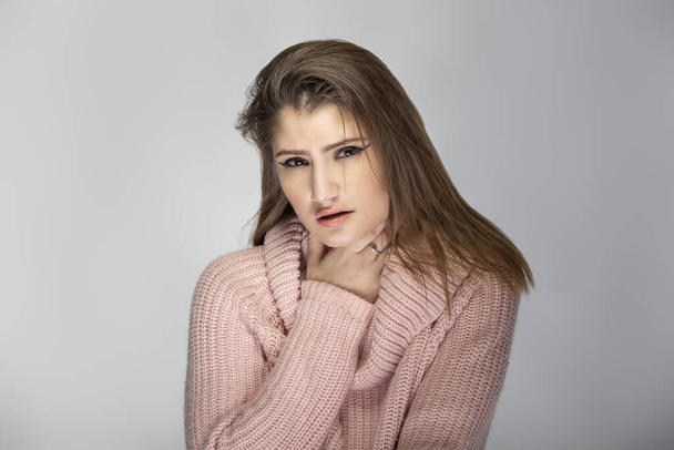 Close up portrait of a young Caucasian woman wearing a pink sweater on a grey background.  The model looks sick with a sore throat - 写真・画像