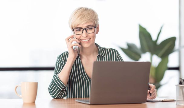 Женщина разговаривает по мобильному телефону с помощью ноутбука, работающего в офисе
 - Фото, изображение