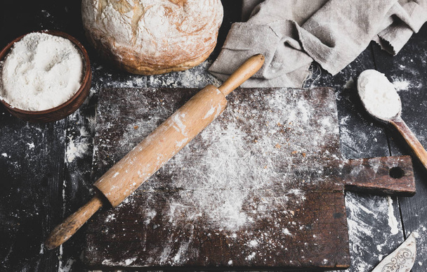 baked bread, white wheat flour, wooden rolling pin and old cutting board on a black table, top view - Фото, изображение