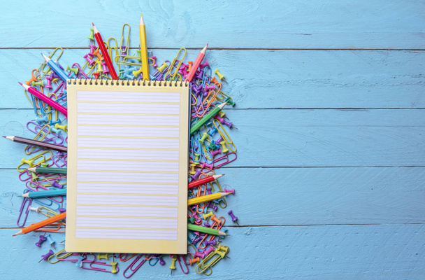 Blank page spiral notebook on a pile of office supplies, colored paper clamps, push pins and wooden crayons. - 写真・画像