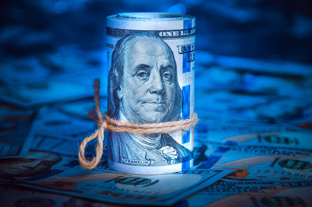 A roll of dollars on the background of scattered hundred dollar bills in blue light - Photo, Image