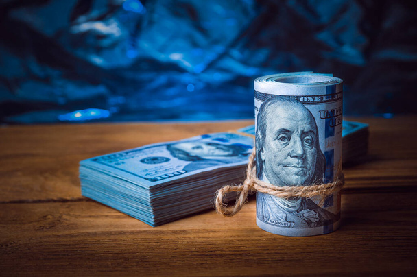 A roll of dollars with a pack of dollars on the background of textured wooden boards in blue light - Photo, Image
