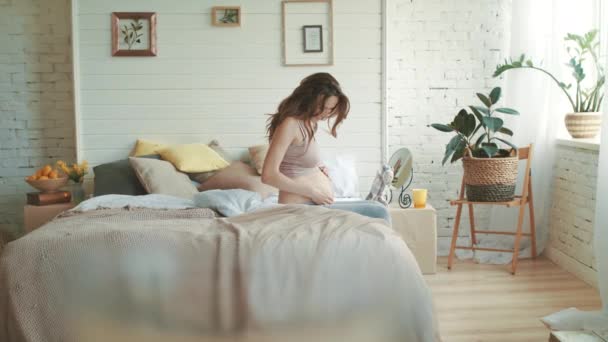 Young pregnant woman sitting in bed at home. Expecting mother touching belly. - Felvétel, videó