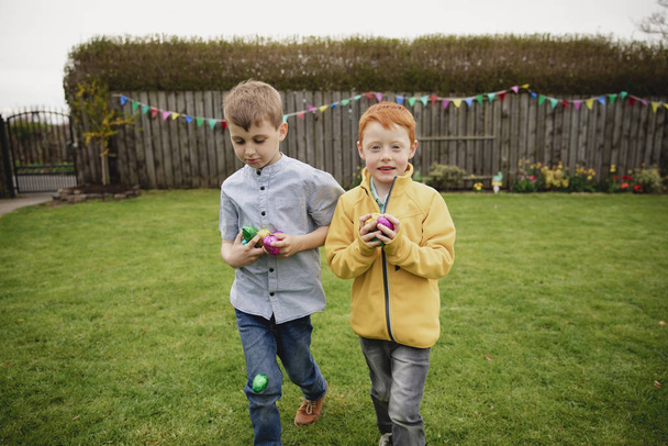 Two young boys walking through the back garden after an easter egg hunt. They are holding handfuls of chocolate eggs in their hands. - Foto, imagen