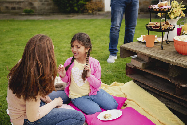 Little girl and her mother sitting outdoors on a blanket while talking. The little girl is talking and playing with her braided hair while at a garden party. - Photo, Image