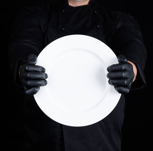 Chef in black uniform and black latex gloves holding a round empty plate - Fotó, kép