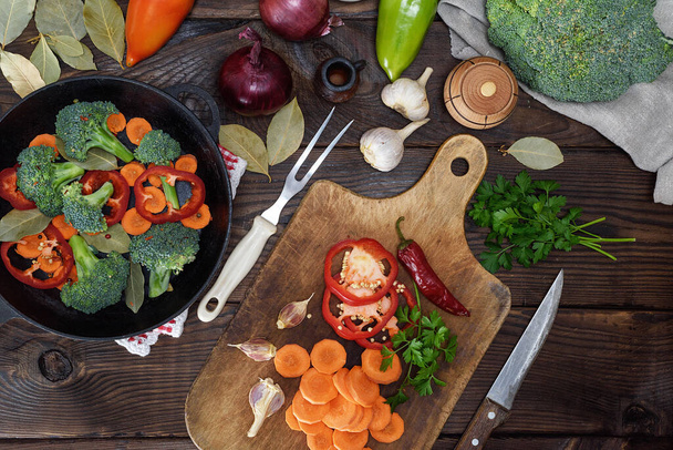 fresh pieces of carrots, broccoli and red pepper on a wooden kitchen board, next to a round cast-iron frying pan - Photo, image