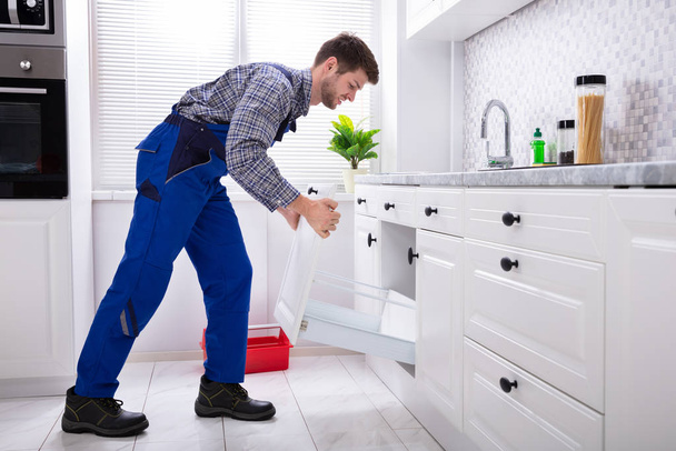 Side View Of A Young Handyman In Uniform Installing Drawer In Kitchen - 写真・画像