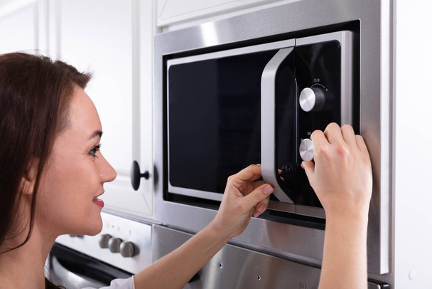Side View Of A Young Woman Using Microwave Oven In Kitchen - Foto, imagen