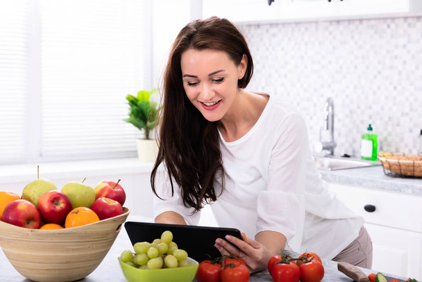 Mujer joven feliz usando tableta digital cerca de verduras frescas en el mostrador de cocina
 - Foto, Imagen