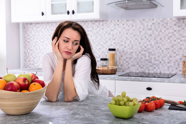 Fresh Fruits In Front Of Upset Young Woman Leaning On Kitchen Counter - Fotografie, Obrázek