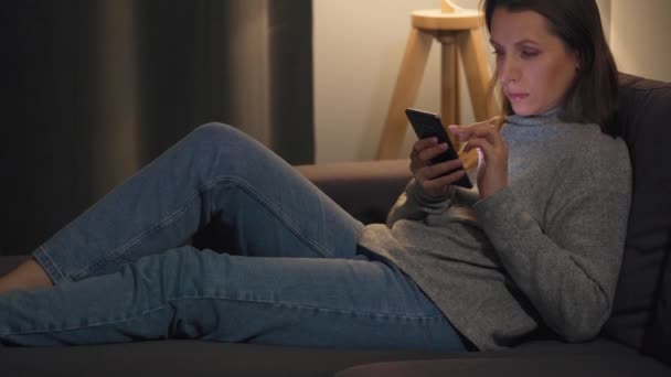 Smiling woman lying on the couch in a cozy room and using smartphone for surfing internet in the evening. Relaxation and lifestyle technology. - Materiaali, video