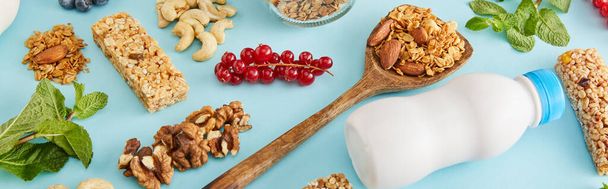 Пищевой состав орехов, бутылка йогурта, ягоды, батончики и мята на голубом фоне, панорамный снимок
 - Фото, изображение