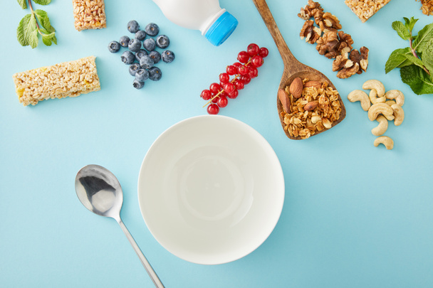 Top view of empty bowl, spoon, berries, spatula, mint, nuts and cereal bars on blue background - Фото, изображение
