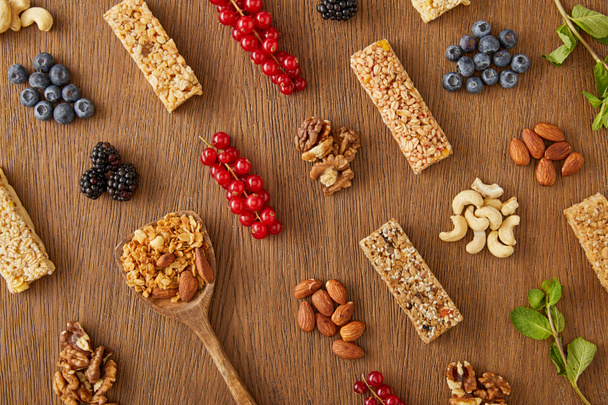 Top view of food composition of berries, nuts, cereal bars, mint and spatula with granola on wooden background - Фото, зображення