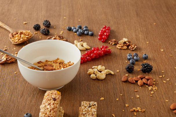 Cuenco con granola junto a bayas, frutos secos y barras de cereales sobre fondo de madera
 - Foto, imagen