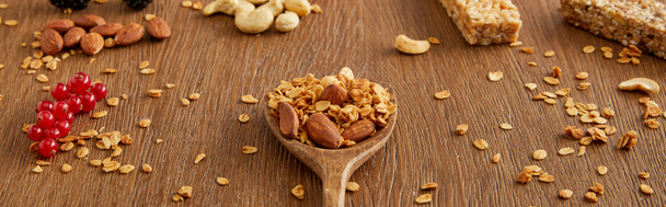 Wooden spatula with granola next to redcurrants, nuts and cereal bars on wooden background, panoramic shot - Fotó, kép