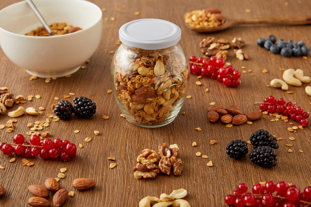 Selective focus of jar of granola and bowl next to nuts, oat flakes and berries on wooden background - Fotó, kép