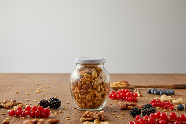Selectieve focus van kruik granola met noten, havervlokken, bessen en graanrepen op houten ondergrond geïsoleerd op grijze ondergrond - Foto, afbeelding