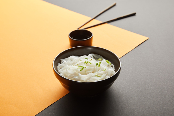 селективный фокус рисовой лапши в миске возле палочек для еды и соевого соуса на желтой и черной поверхности
 - Фото, изображение