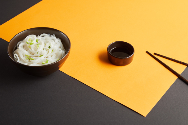 rýžové nudle v misce poblíž hůlek a sójové omáčky na žlutém a černém povrchu - Fotografie, Obrázek