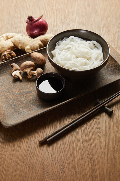 рисовая лапша в миске возле палочек, соевый соус, корень имбиря, лук и грибы на деревянном подносе
 - Фото, изображение