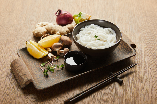 rice noodles in bowl near chopsticks, soy sauce, ginger root, lemon and vegetables on wooden tray - Foto, immagini