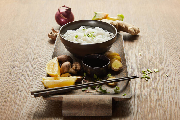 rice noodles in bowl near chopsticks, soy sauce, ginger root, lemon and vegetables on wooden tray - Foto, afbeelding