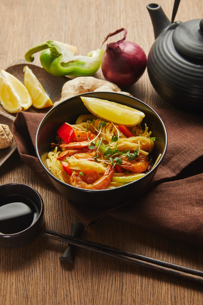 noodles with shrimps and vegetables in bowl near chopsticks, soy sauce, lemon and ginger root, teapot on napkin on wooden table - Photo, Image