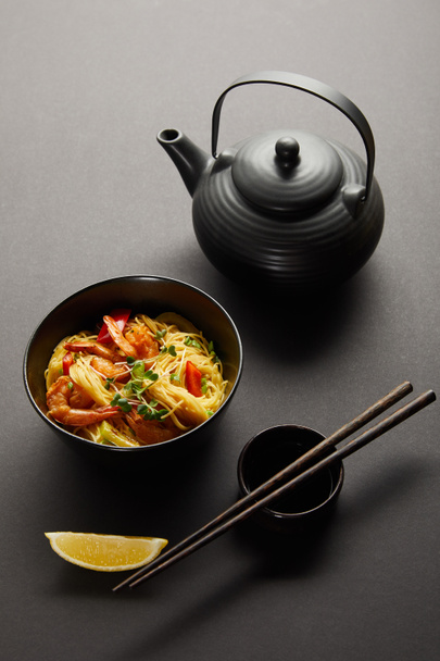noodles with shrimps and vegetables in bowl near lemon, teapot, wooden chopsticks and soy sauce on black background - Photo, Image