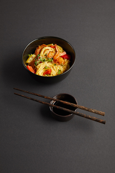noodles with shrimps and vegetables in bowl near wooden chopsticks and soy sauce on black background - Photo, Image