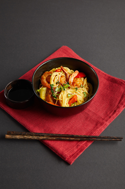 noodles with shrimps and vegetables in bowl near wooden chopsticks, soy sauce on red napkin on black background - Foto, afbeelding