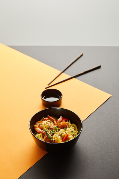 selektivní zaměření nudlí s krevetami a zeleninou v misce v blízkosti hůlek, sójová omáčka na černém a žlutém pozadí izolované na šedé - Fotografie, Obrázek