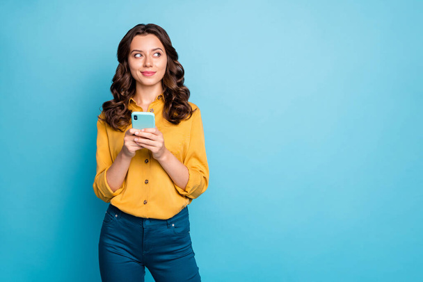 Photo of pretty wavy lady holding telephone hands thinking over creative post text idea looking side empty space wear yellow shirt trousers isolated blue color background - Φωτογραφία, εικόνα