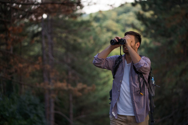 Happy adult man is hiking in forest. He is using binoculars. - Photo, Image