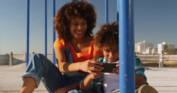 Front view of mixed race woman with her son enjoying family time at the playground together on a sunny day with blue sky, using a smartphone, smiling, slow motion - Metraje, vídeo