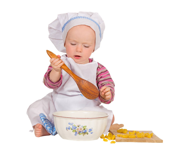 Cute young baby dressed in an apron and chef's toque sitting on the floor with a bowl, wooden spoon and homemade biscuits learning to bake - Foto, immagini