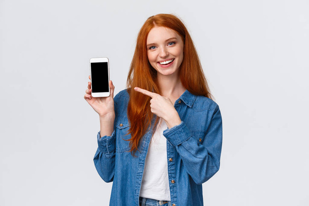 Pas-up portret radosny charyzmatyczny ruda kobieta recenzja smartphone aplikacji, pokazując jej zdjęcie z cute filtr, wskazując mobilny wyświetlacz i uśmiechnięty aparat fotograficzny, białe tło - Zdjęcie, obraz