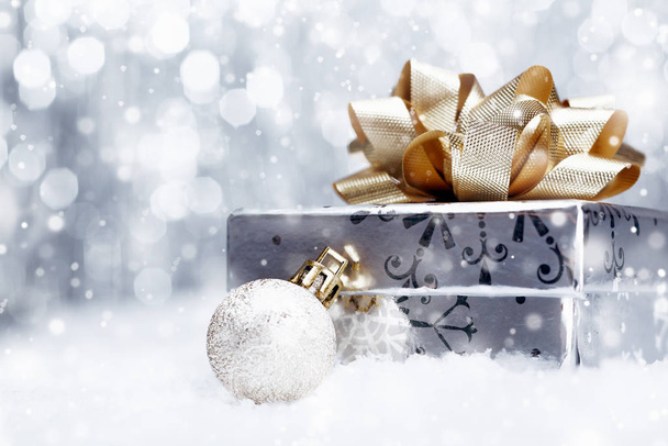 Silver Christmas gift with a huge ornamental golden bow lying with a bauble on a bed of fresh snow with falling snowflakes and copyspace - Foto, Imagem
