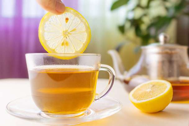 Un limón se coloca en una taza de té a base de plantas. taza transparente. En el fondo es un teapot. El concepto de bebidas orgánicas saludables. - Foto, Imagen
