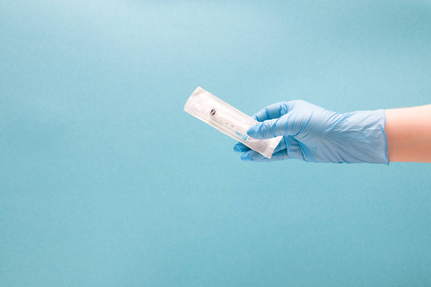 vrouwelijke hand in een blauwe wegwerp medische handschoen houdt een wegwerpspuit in een verpakking, blauwe achtergrond kopieerruimte - Foto, afbeelding