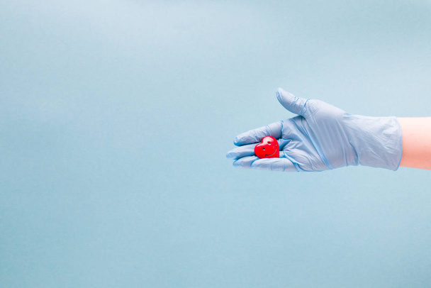 a female hand in a blue disposable medical glove holds a small red heart in her palm with a smile, a smiling heart, health care concept, blue background, copy space, varnish cordiologist cares about heart health - Fotoğraf, Görsel