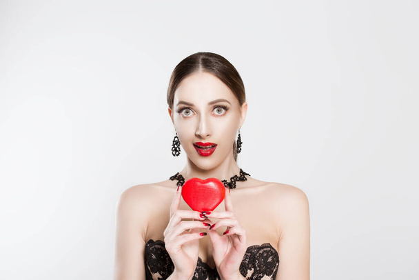 The girl showing the affection, holds the heart in her hands, the expression of the girl is a little surprise, amazing isolated white background. Red heart box, lips and black dress earrings chokers - Zdjęcie, obraz