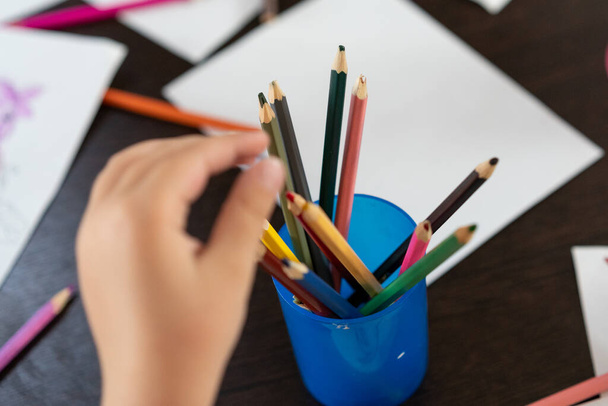 colored pencils in a glass. Behind the pencils is a child's hand. A small child wants to draw. Colored pencils in disorder to lie on the table. - Photo, image