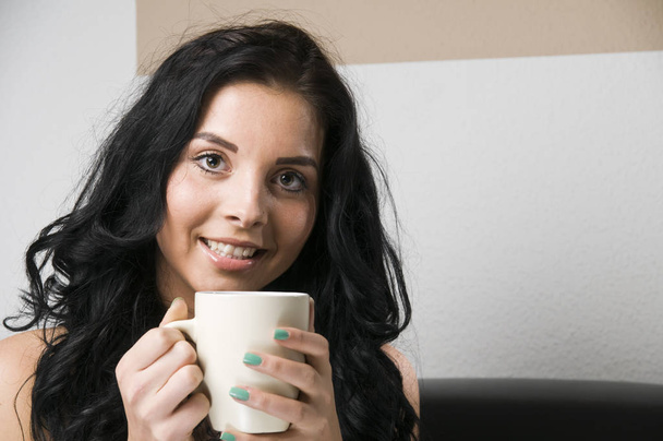 head-and-shoulder-shot of a young black-haired woman with white cup in hands and smiling at the camera - Fotografie, Obrázek