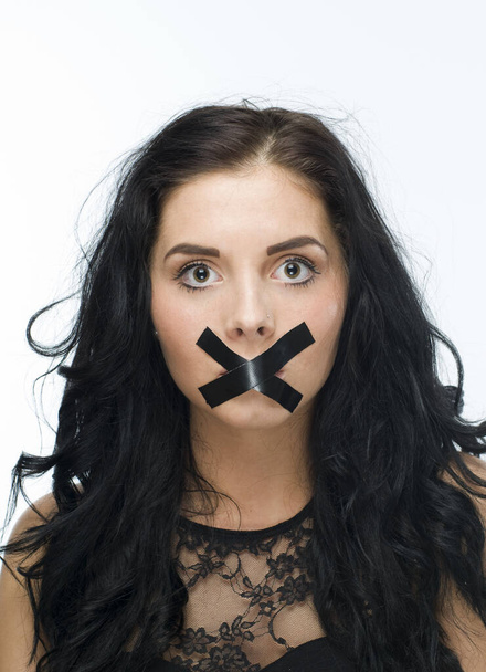 head-and-shoulder-shot of a young black-haired woman against white background and with black tape crosswise glued mouth - 写真・画像